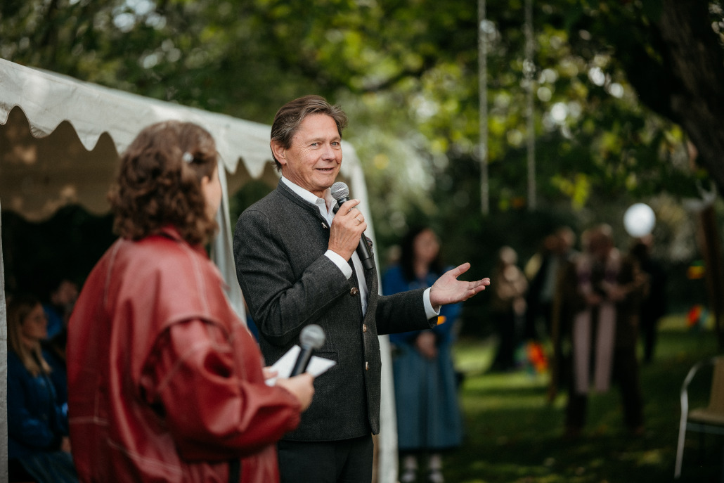 Beim Eröffnungsfest St. Veit spricht ein Mann im Anzug in ein Mikrofon im Freien, während eine andere Person mit einem Mikrofon in der Nähe steht; im Hintergrund versammeln sich Menschen unter Bäumen und einem Zelt.