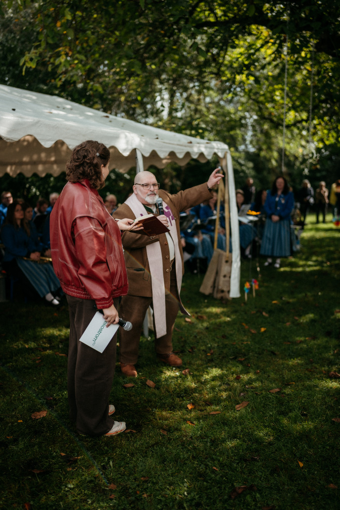 Auf dem Eröffnungsfest St. Veit spricht und gestikuliert eine Person in Anzug und Stola neben einer anderen Person in roter Jacke bei einer Veranstaltung im Freien unter einem weißen Zelt, während im Hintergrund Menschen sitzen.