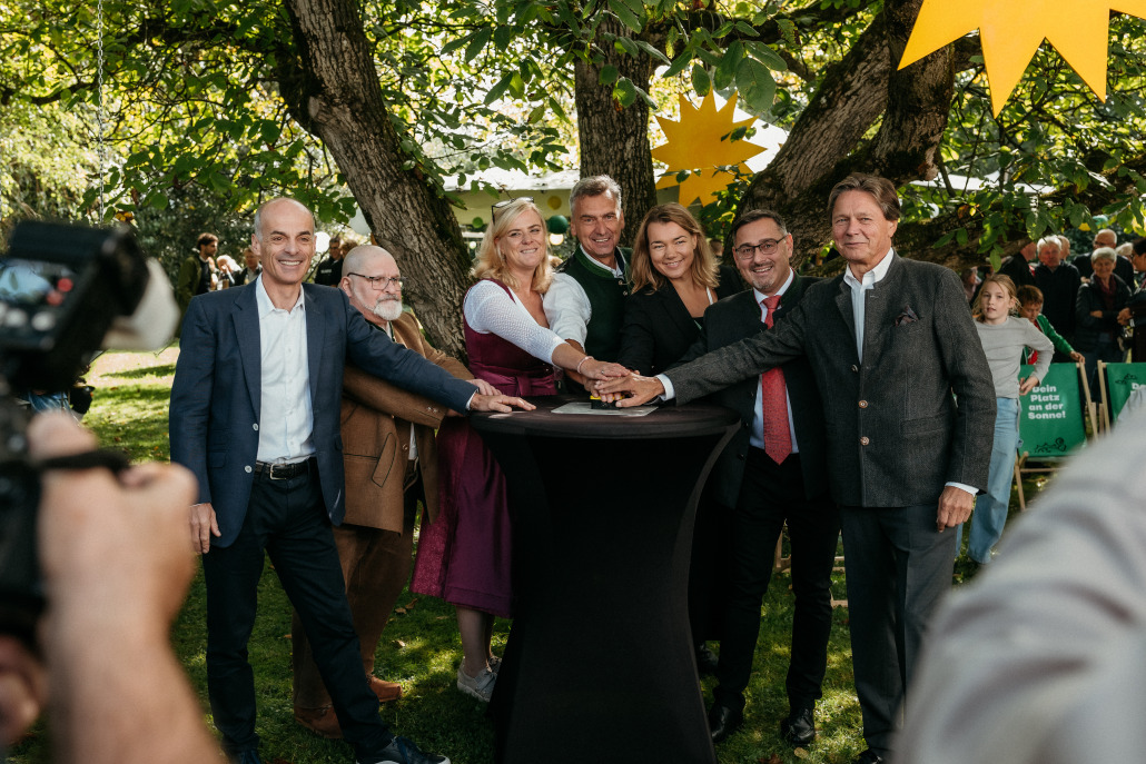 Sieben Erwachsene stehen im Freien um einen hohen schwarzen Tisch, jeder mit einer Hand auf dem Tisch, und posieren für ein Gruppenfoto während des Eröffnungsfestes St. Veit.