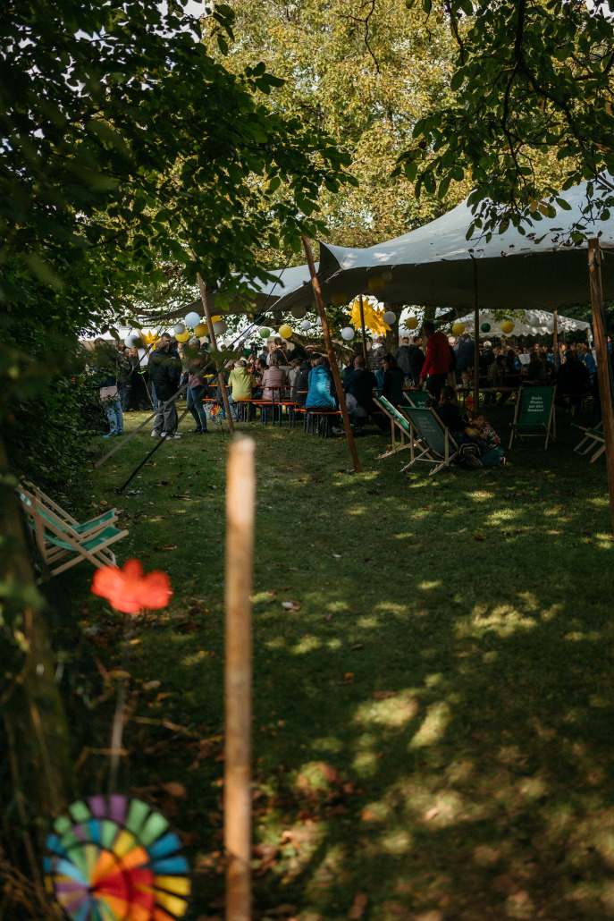 Beim Eröffnungsfest St. Veit versammeln sich die Menschen unter einem großen weißen Zelt, das mit gelben Luftballons geschmückt ist. Liegestühle und Tische sind auf dem grasbewachsenen, schattigen Gelände aufgestellt, im Vordergrund stehen Bäume und ein buntes Windrad.