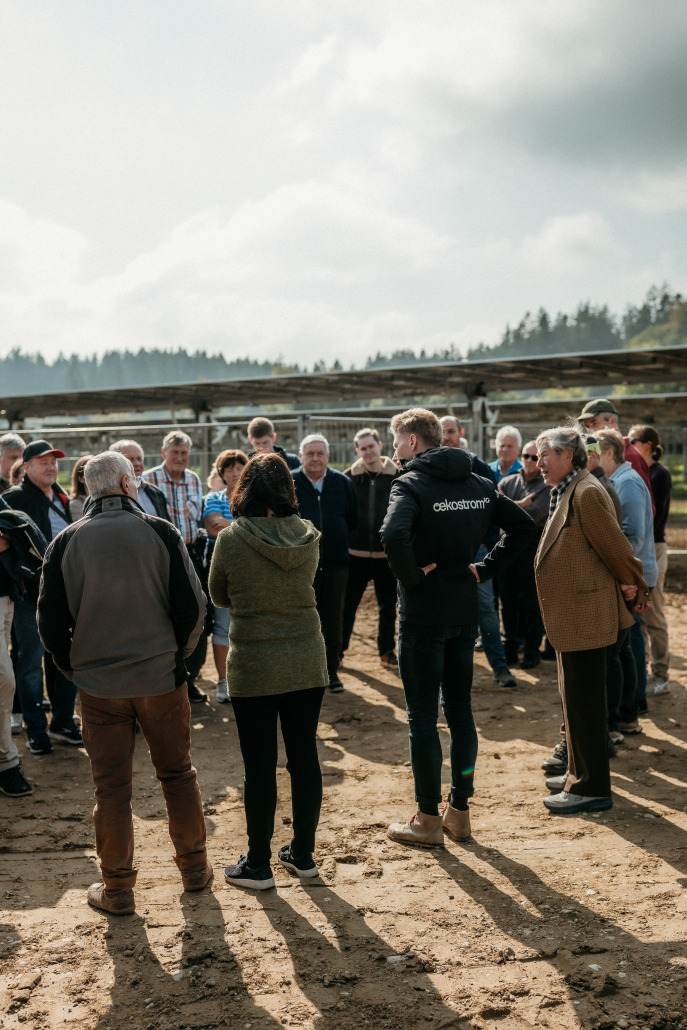 Eine Gruppe von Menschen steht im Kreis auf einer unbefestigten Fläche im Freien und diskutiert unter einem wolkenverhangenen Himmel mit Bäumen im Hintergrund beim Eröffnungsfest St. Veit.