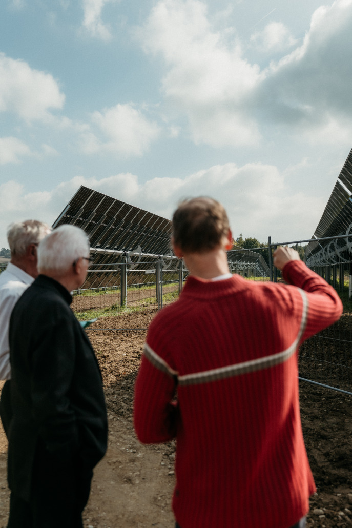 Drei Personen stehen beim Eröffnungsfest in St. Veit an einem Zaun und beobachten große Solarpaneele im Freien unter einem teilweise bewölkten Himmel.