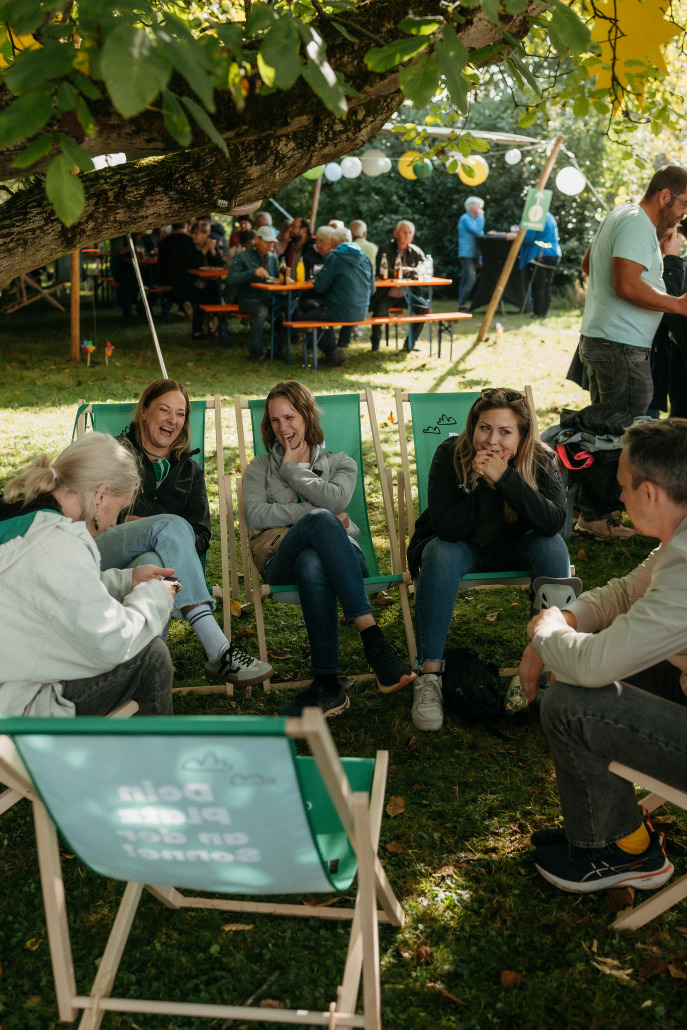Eine Gruppe von Menschen sitzt auf Liegestühlen und unterhält sich unter einem Baum auf dem Eröffnungsfest St. Veit, während andere an Picknicktischen im Hintergrund Platz nehmen.