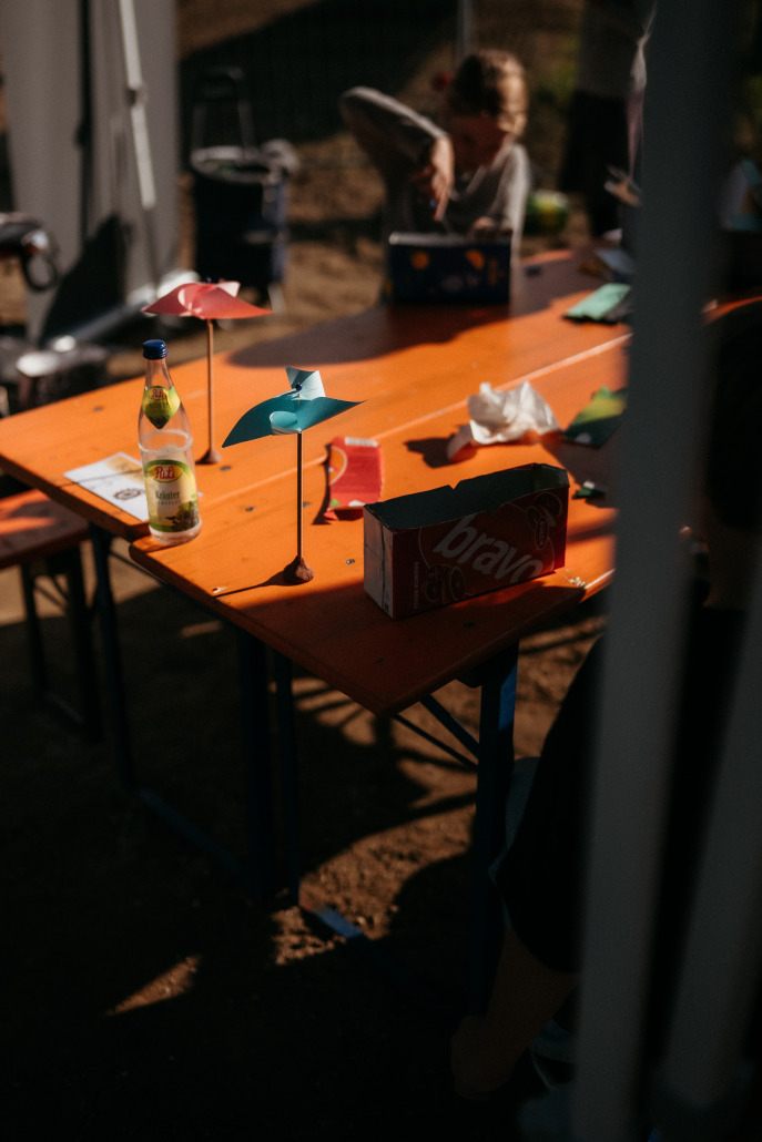 Ein leuchtend orangefarbener Tisch im Freien mit Papierwindrädern, einer Flasche Zitronen-Eistee, einer Dose Bravo-Fruchtsaft, Taschentüchern und mehreren Personen, die teilweise um ihn herum zu sehen sind, beim Eröffnungsfest St. Veit.