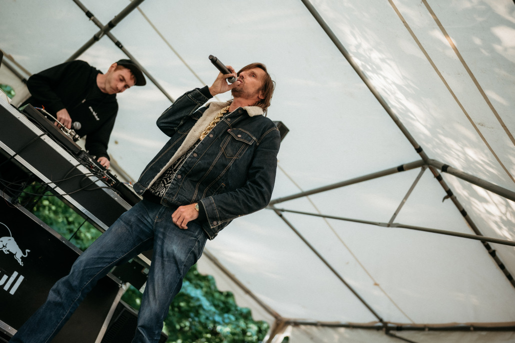 Ein Mann singt in ein Mikrofon, während eine andere Person unter einem weißen Baldachin beim Eröffnungsfest St. Veit eine DJ-Anlage bedient.