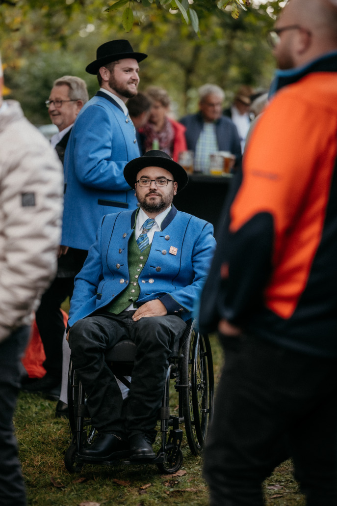 Ein Mann im Rollstuhl, der die traditionelle blaue Jacke und den schwarzen Hut trägt, sitzt beim Eröffnungsfest St. Veit unter freiem Himmel.