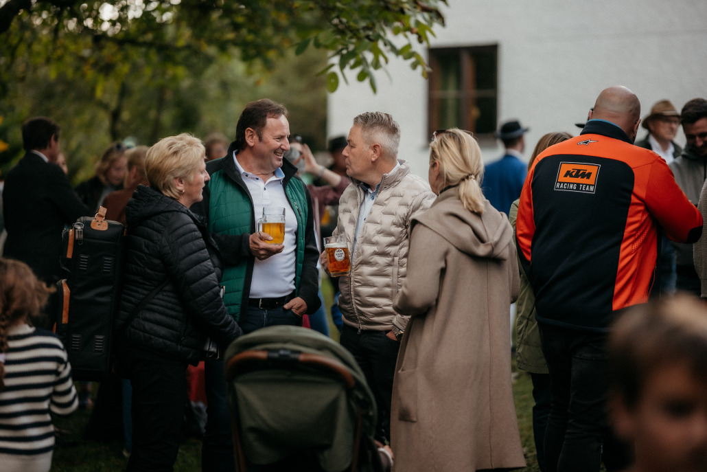 Eine Gruppe Erwachsener steht beim Eröffnungsfest St. Veit im Freien, unterhält sich und hält Getränke in der Hand, während im Hintergrund andere Personen und ein Kinderwagen zu sehen sind.