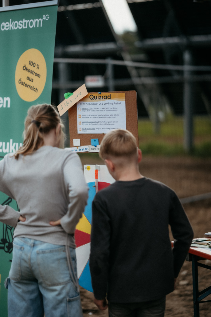Zwei Kinder stehen beim Eröffnungsfest St. Veit vor einem Quizrad und lesen die Informationen und Regeln auf der Tafel während der lebhaften Veranstaltung im Freien.