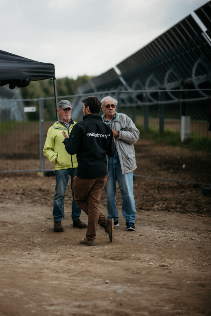 Drei Männer stehen beim Eröffnungsfest in St. Veit in der Nähe eines eingezäunten Geländes mit großen Solarpanels und unterhalten sich; einer trägt eine Jacke der Marke Celestron.