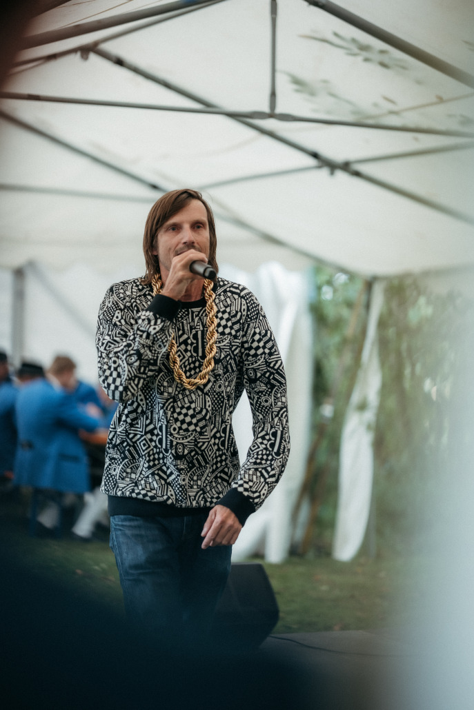 Ein Mann mit einem gemusterten Pullover und Goldketten hält ein Mikrofon in der Hand und tritt unter einem weißen Zelt auf dem Eröffnungsfest St. Veit auf.