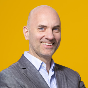 Ein glatzköpfiger Mann in grauem Anzug und hellblauem Hemd lächelt in die Kamera vor einem einfarbig gelben Hintergrund, der den professionellen Geist der Arbeitsplätze bei der oekostrom AG widerspiegelt.