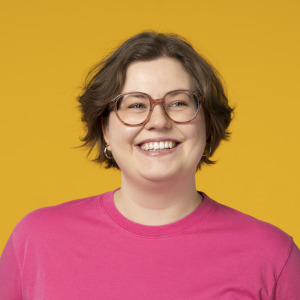 Eine Person mit kurzen braunen Haaren, Brille und rosa Hemd lächelt vor einem einfarbig gelben Hintergrund und verkörpert die positive Energie der jobs oekostrom AG.