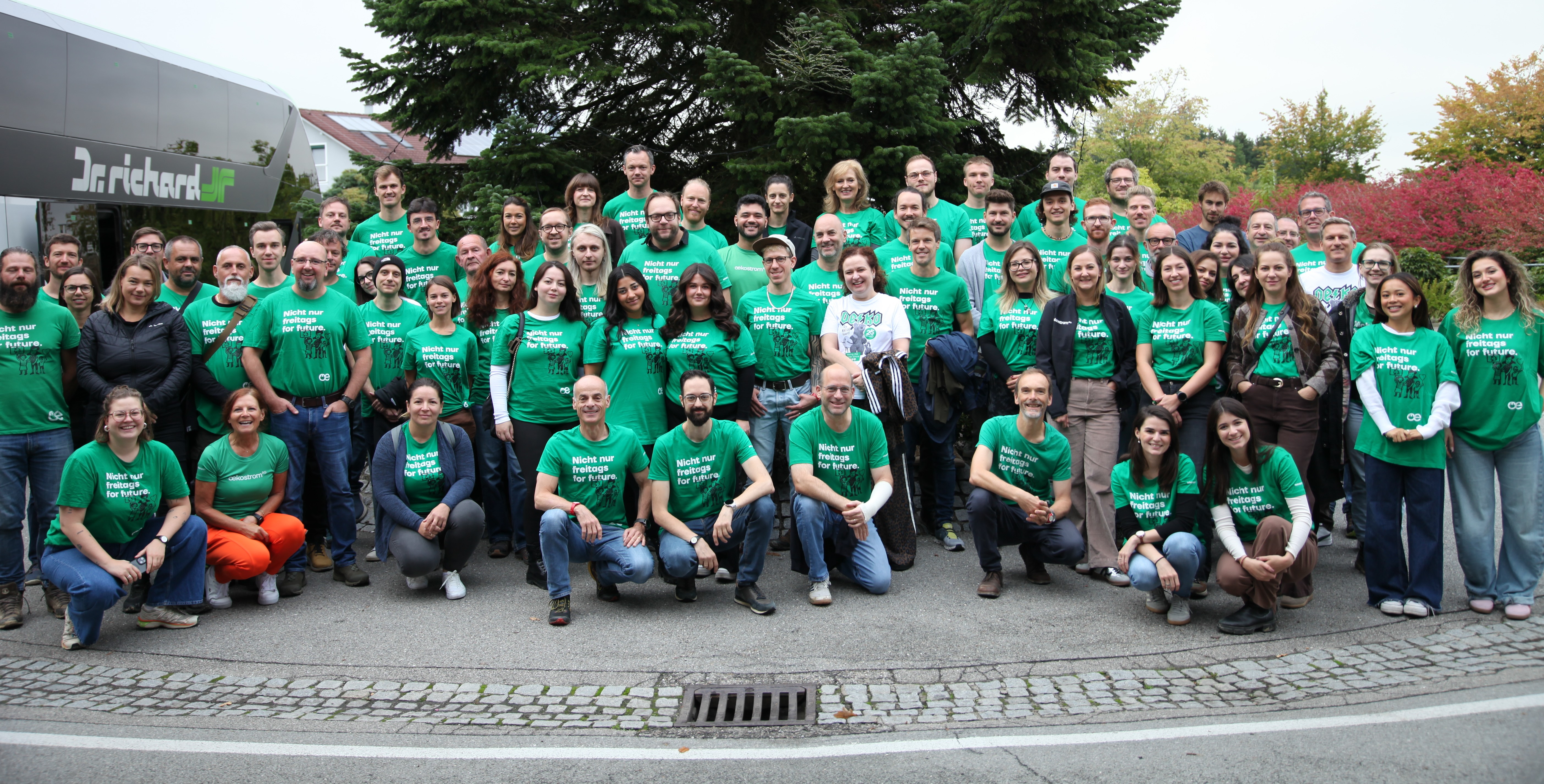 Eine große Gruppe von Menschen in passenden grünen T-Shirts, die die jobs oekostrom AG repräsentieren, posieren für ein Gruppenfoto im Freien auf einer gepflasterten Fläche neben Bäumen und einem Gebäude.