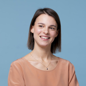 Eine Frau mit glattem braunem Haar, die eine pfirsichfarbene Bluse und eine Halskette trägt, lächelt, während sie vor einem schlichten blauen Hintergrund leicht zur Seite schaut - perfekt für die Darstellung von Fachleuten bei der oekostrom AG.