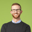 Mann mit kurzen roten Haaren, Bart und Brille, der einen dunklen Pullover über einem Kragenhemd trägt, lächelt, während er vor einem grünen Hintergrund steht.