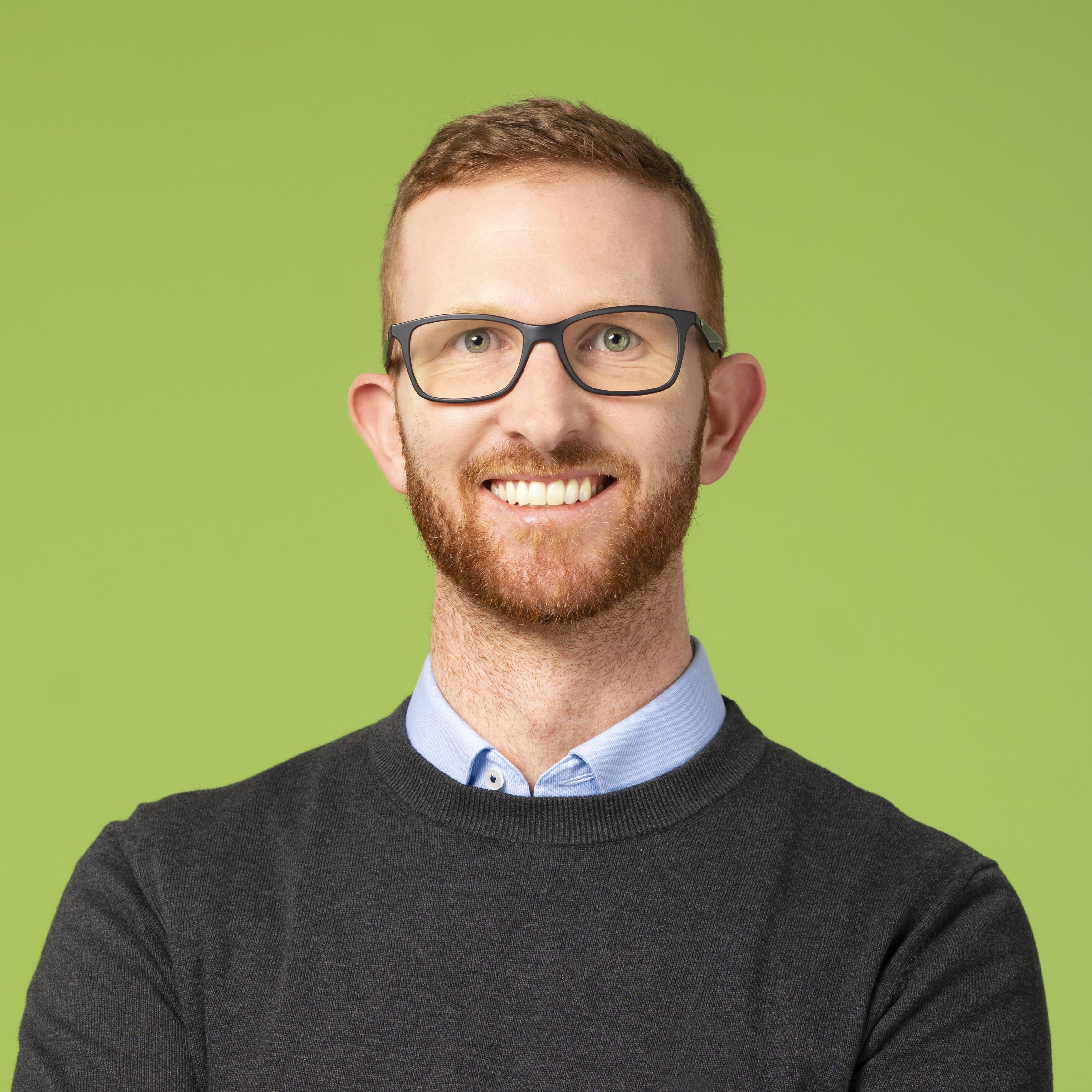 Mann mit kurzen roten Haaren, Bart und Brille, der einen dunklen Pullover über einem Kragenhemd trägt, lächelt, während er vor einem grünen Hintergrund steht.