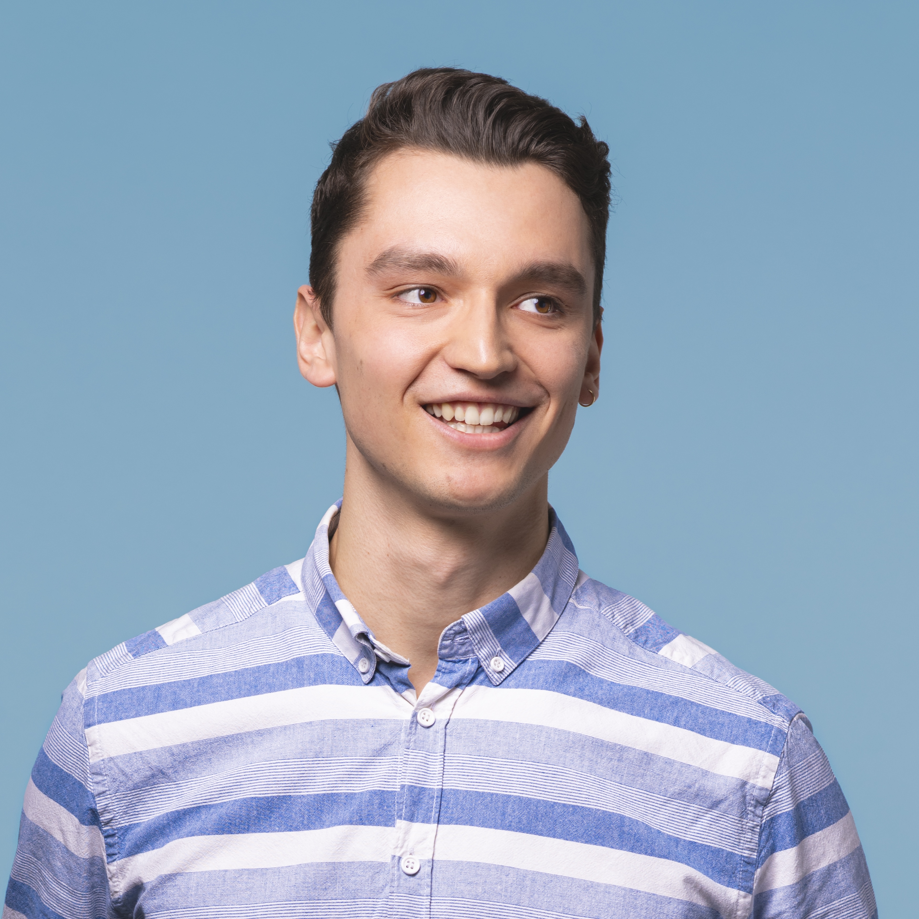 Ein junger Mann in einem blau-weiß gestreiften Hemd lächelt und blickt vor einem einfarbig hellblauen Hintergrund zur Seite.