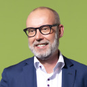 Ein lächelnder Mann mit Bart und Brille, der ein weißes Hemd und einen blauen Blazer trägt, steht vor einem schlichten grünen Hintergrund.