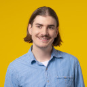 Ein junger Mann mit schulterlangem braunem Haar und Schnurrbart, der ein blau gestreiftes Hemd trägt, lächelt vor einem einfarbig gelben Hintergrund.