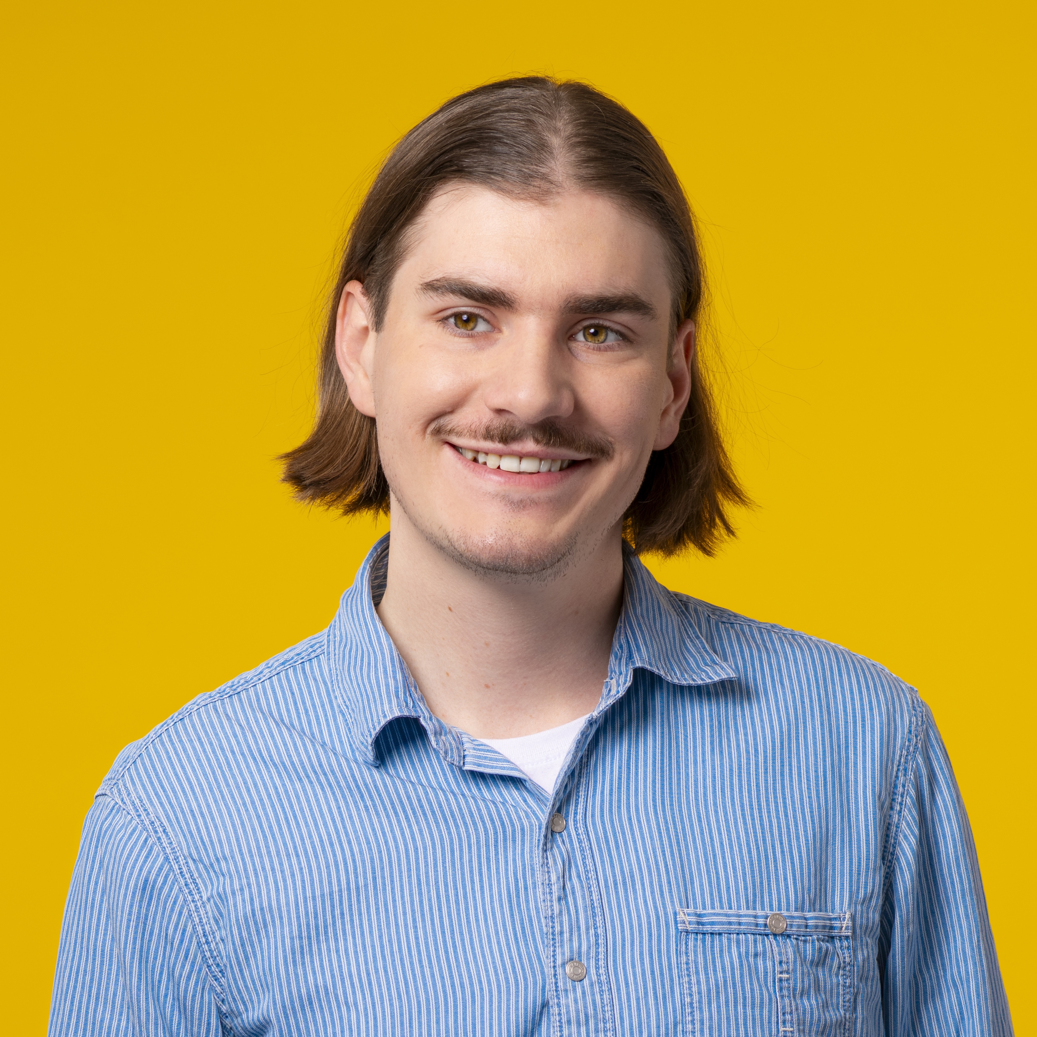 Ein junger Mann mit schulterlangem braunem Haar und Schnurrbart, der ein blau gestreiftes Hemd trägt, lächelt vor einem einfarbig gelben Hintergrund.