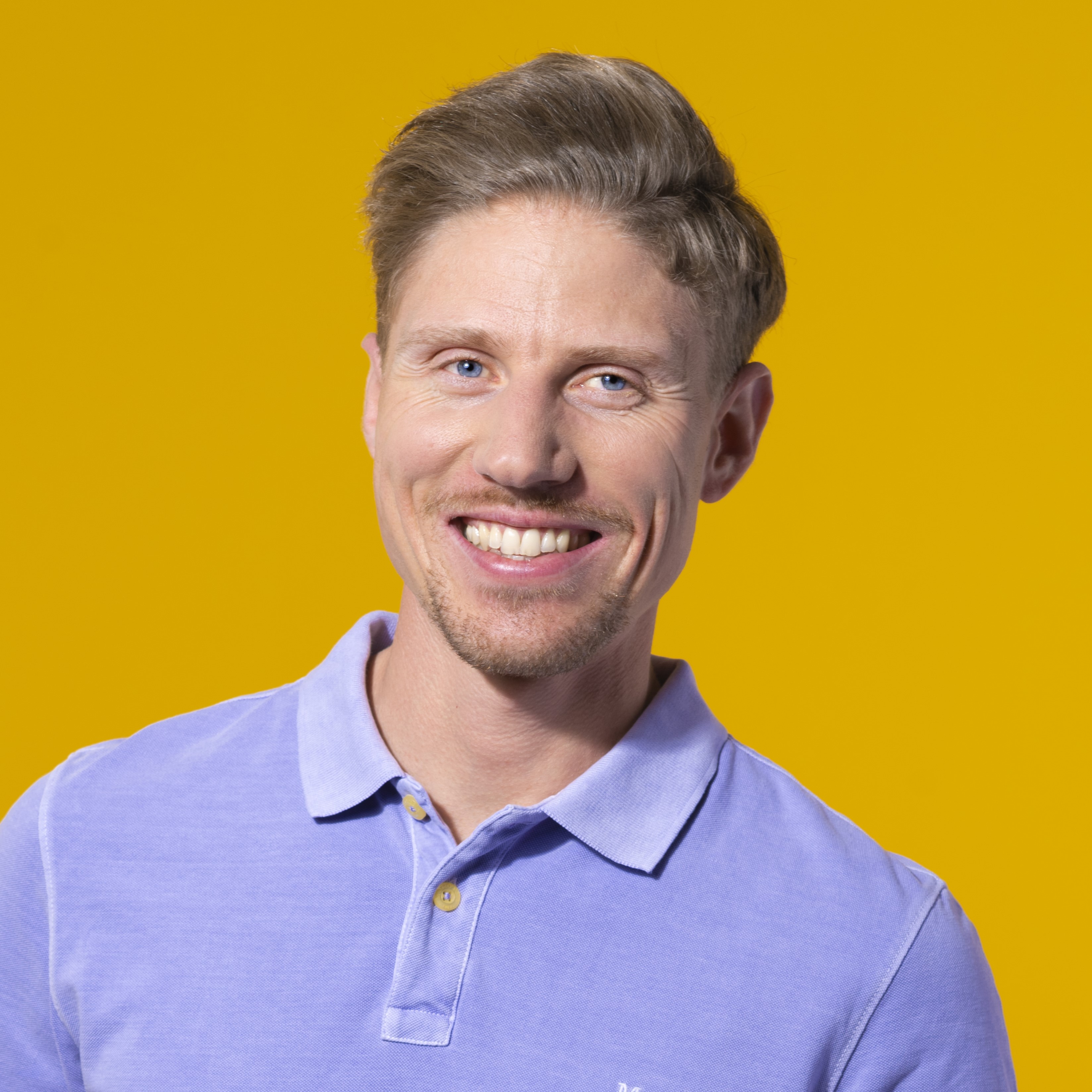 Ein lächelnder Mann mit kurzen hellbraunen Haaren und einem gestutzten Bart trägt ein hellviolettes Polohemd und steht vor einem einfarbig gelben Hintergrund.