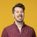 Ein Mann mit kurzen braunen Haaren und gestutztem Bart, der ein rotes Button-up-Hemd trägt, lächelt vor einem schlichten gelben Hintergrund.