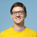 Eine Person mit kurzen braunen Haaren und Brille lächelt, trägt ein gelbes Hemd und steht vor einem schlichten hellblauen Hintergrund.