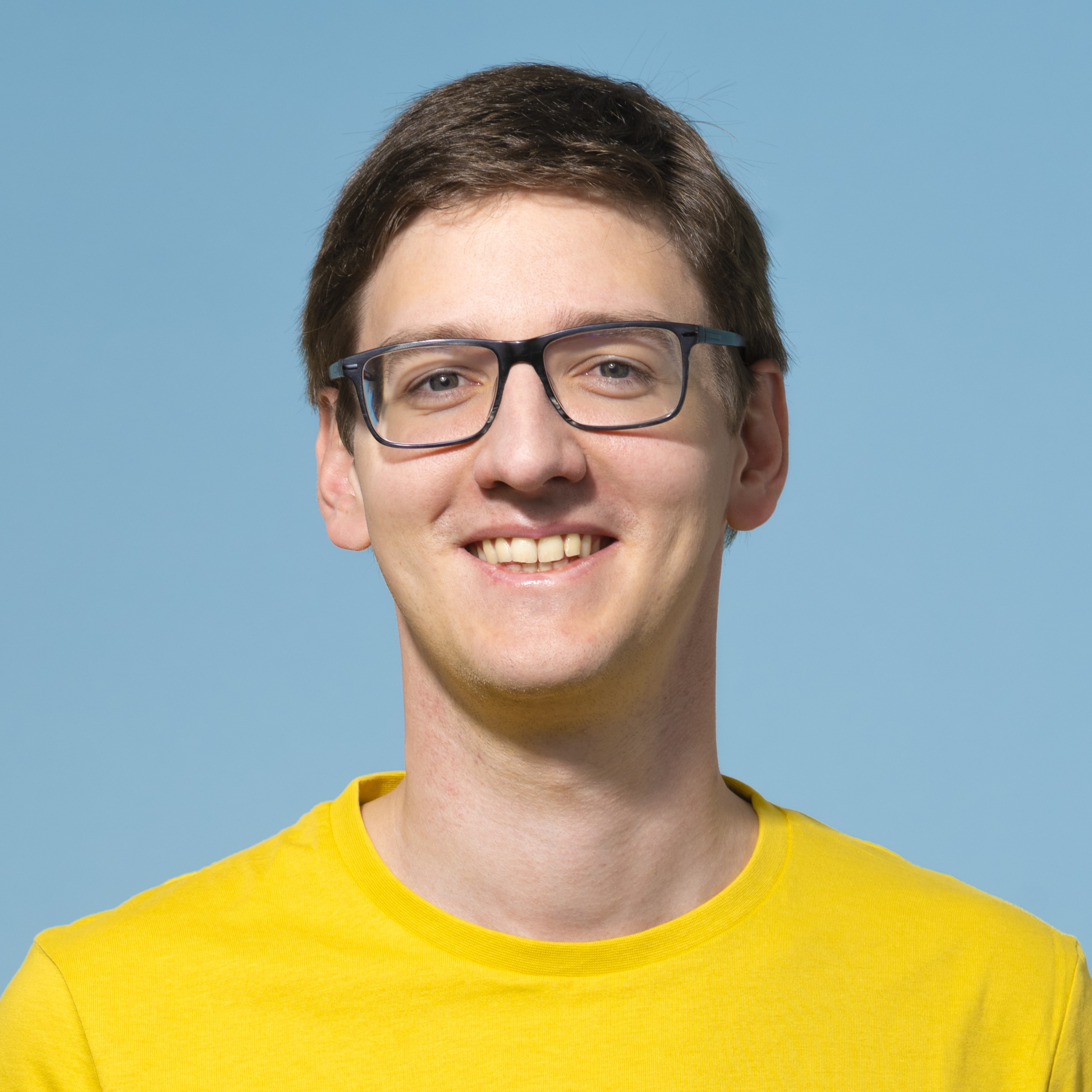 Eine Person mit kurzen braunen Haaren und Brille lächelt, trägt ein gelbes Hemd und steht vor einem schlichten hellblauen Hintergrund.