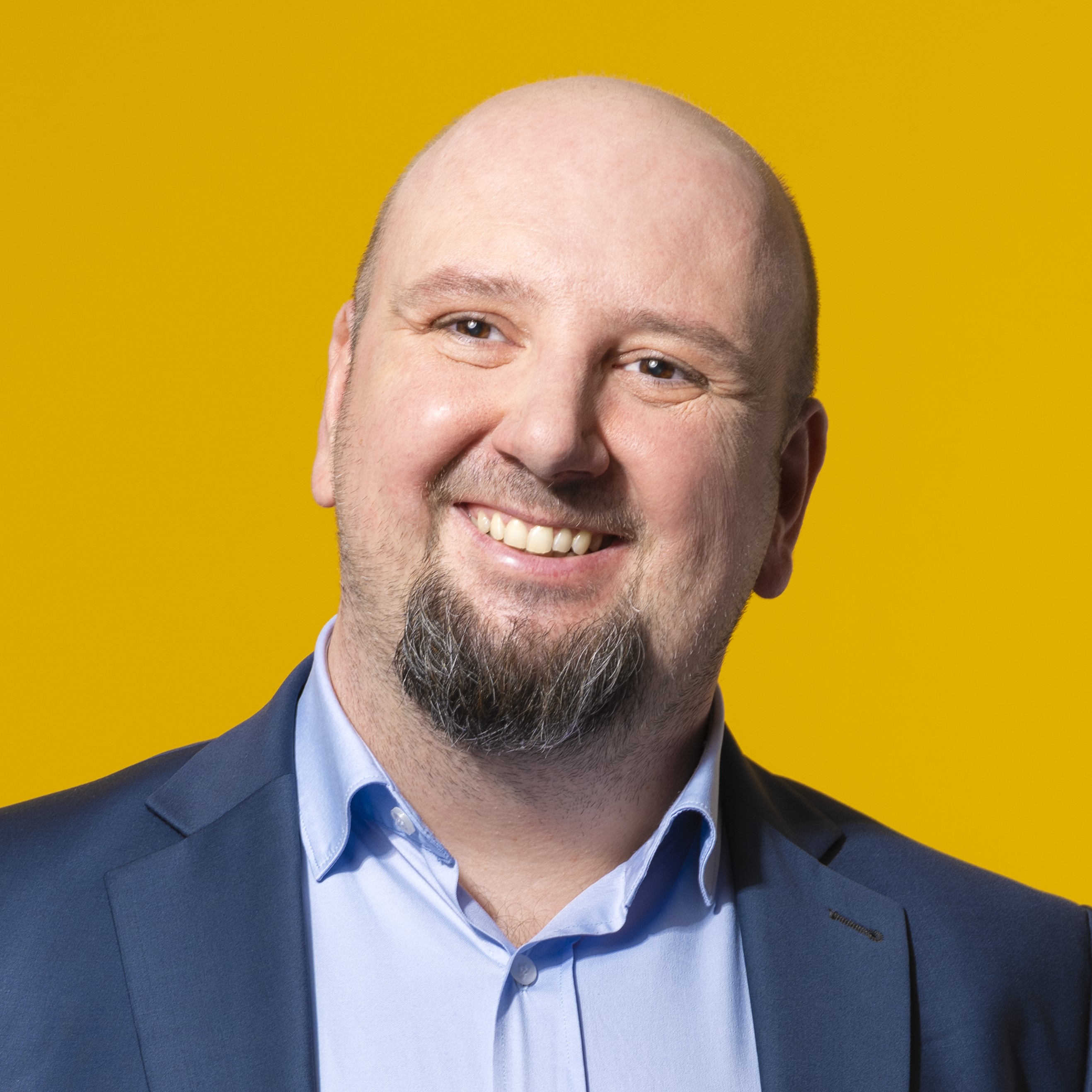 Ein glatzköpfiger Mann mit Bart und Schnurrbart, der eine blaue Anzugsjacke und ein hellblaues Hemd trägt, lächelt vor einem einfarbig gelben Hintergrund.