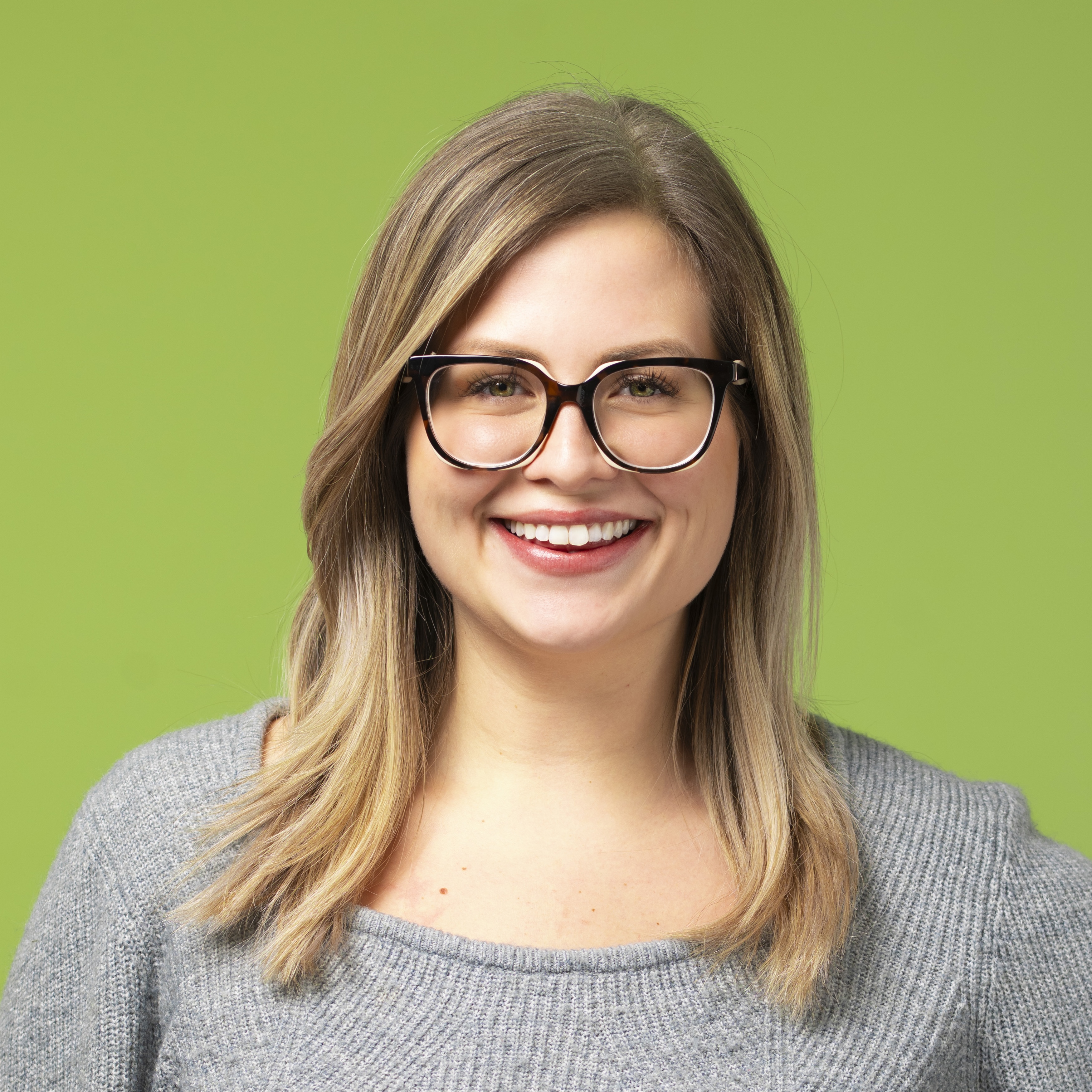 Eine Frau mit langem, hellbraunem Haar und Brille lächelt, trägt einen grauen Pullover und steht vor einem schlichten grünen Hintergrund.