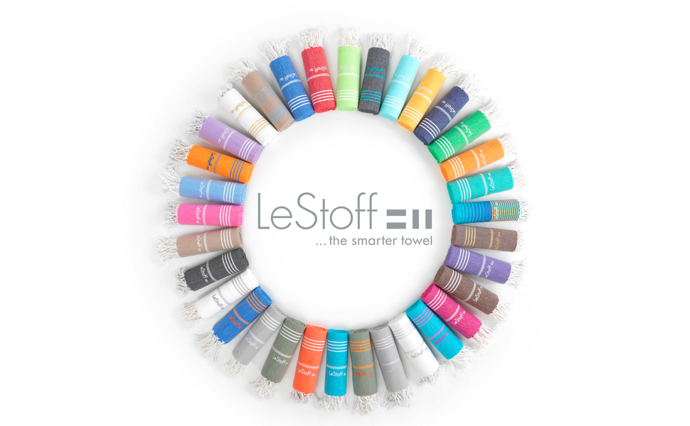 Eine kreisförmige Anordnung von gerollten Handtüchern in verschiedenen Farben umgibt den Text "LeStoff ... the smarter towel" auf weißem Hintergrund.