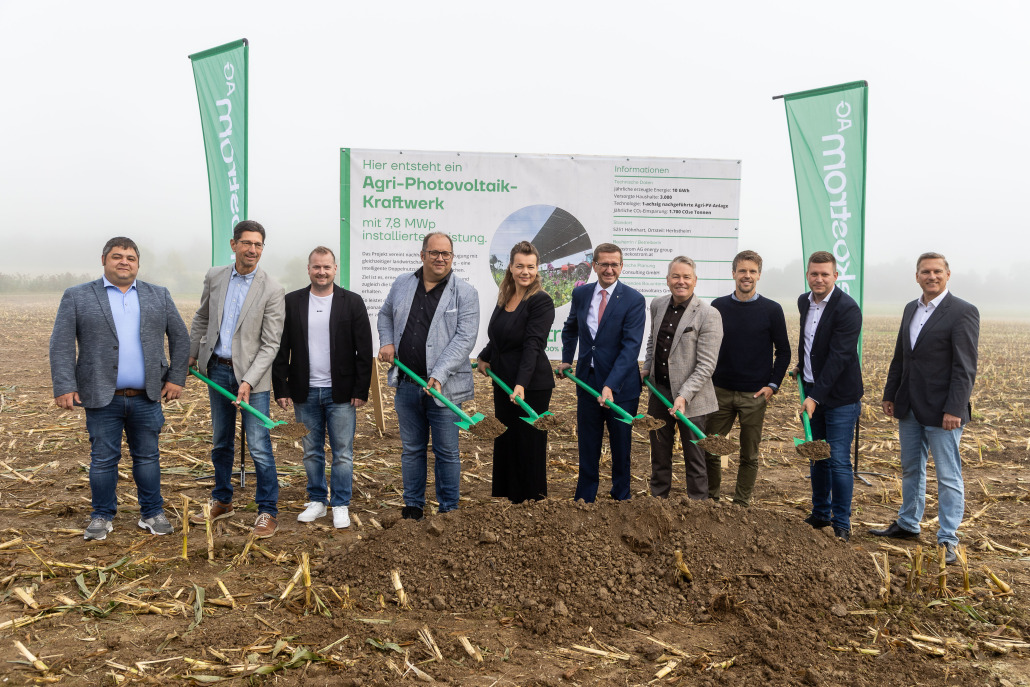 021Oekostrom_AG_Spatenstich Zehn Personen stehen in einer Reihe und halten grüne Schaufeln bei der Grundsteinlegung für ein Photovoltaik-Kraftwerk, im Hintergrund ein Schild und zwei grüne Fahnen.