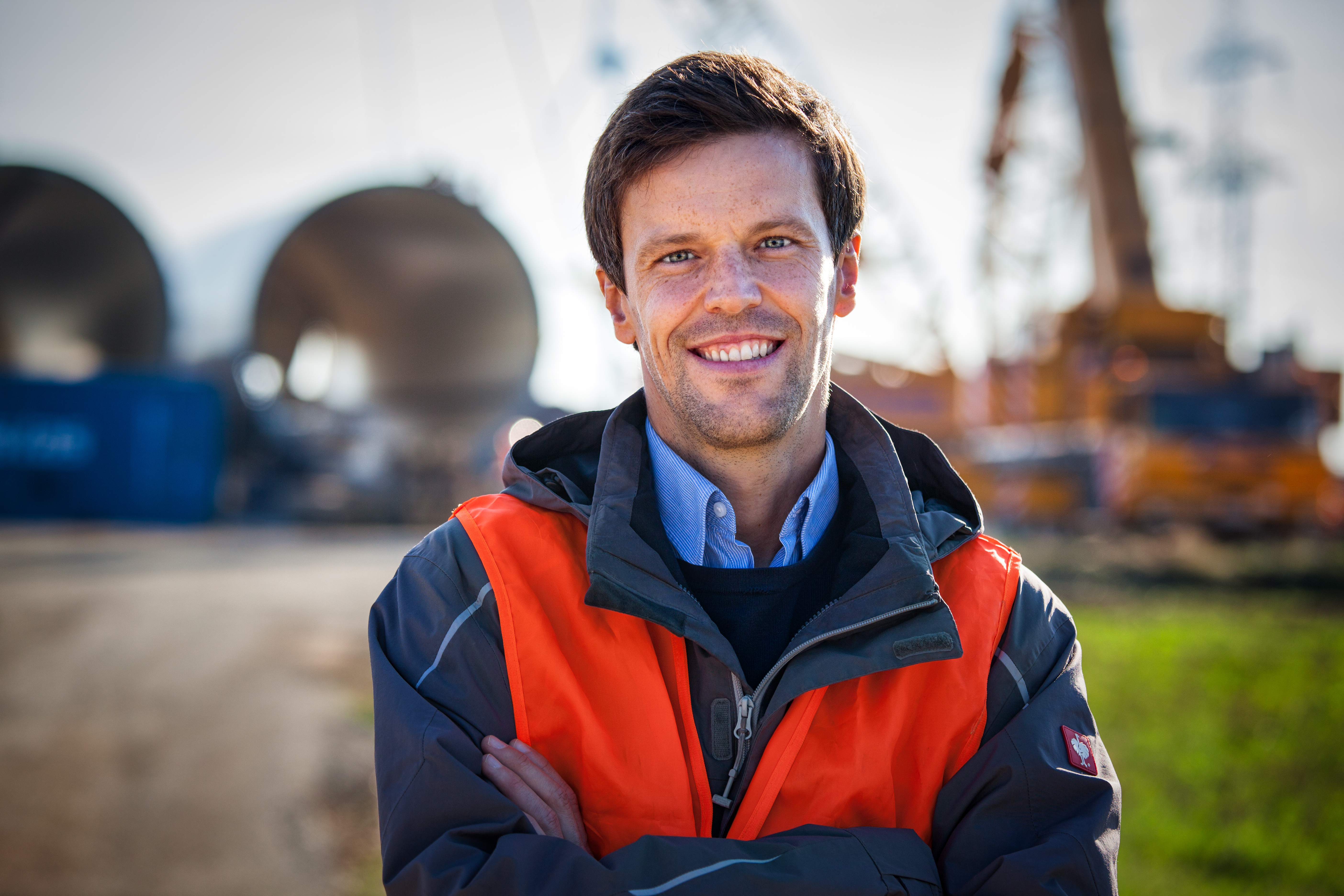 00080_Foto_Thomas_Kirschner_RepParn_Origprop_Max_JEPG_shp_clean Ein Mann mit orangefarbener Sicherheitsweste und Jacke steht im Freien auf einer Baustelle mit großen Strukturen und Geräten im Hintergrund.