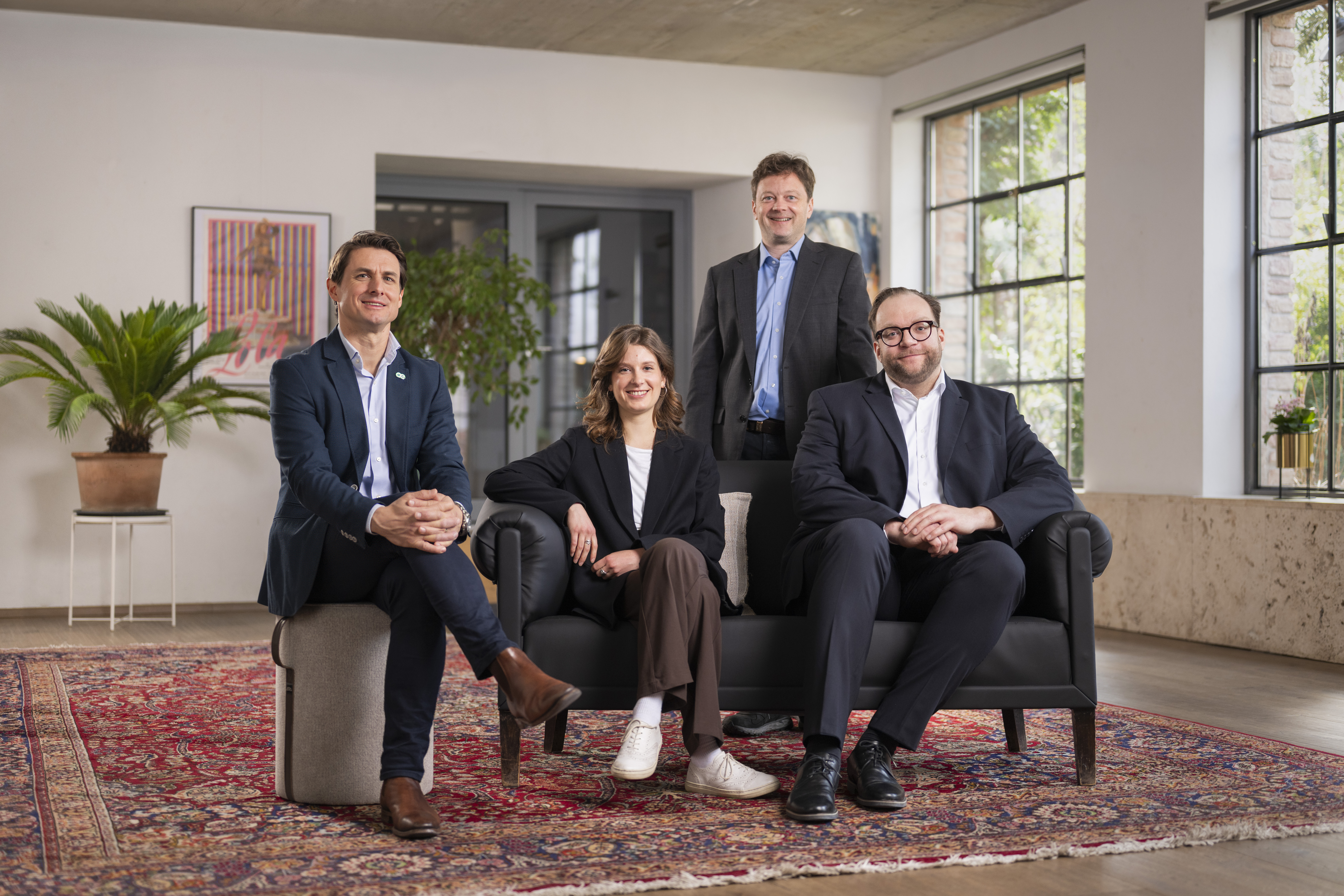 _DSC2327-Bearbeitet-Bearbeitet-Bearbeitet-Bearbeitet-Bearbeitet-2 Vier Personen in Geschäftskleidung posieren in einem modernen, gut beleuchteten Büroraum - vielleicht besprechen sie die Direktvermarktung Kraftwerke - zwei sitzen auf einem schwarzen Sofa, während zwei hinter ihnen auf einem gemusterten Teppich stehen.