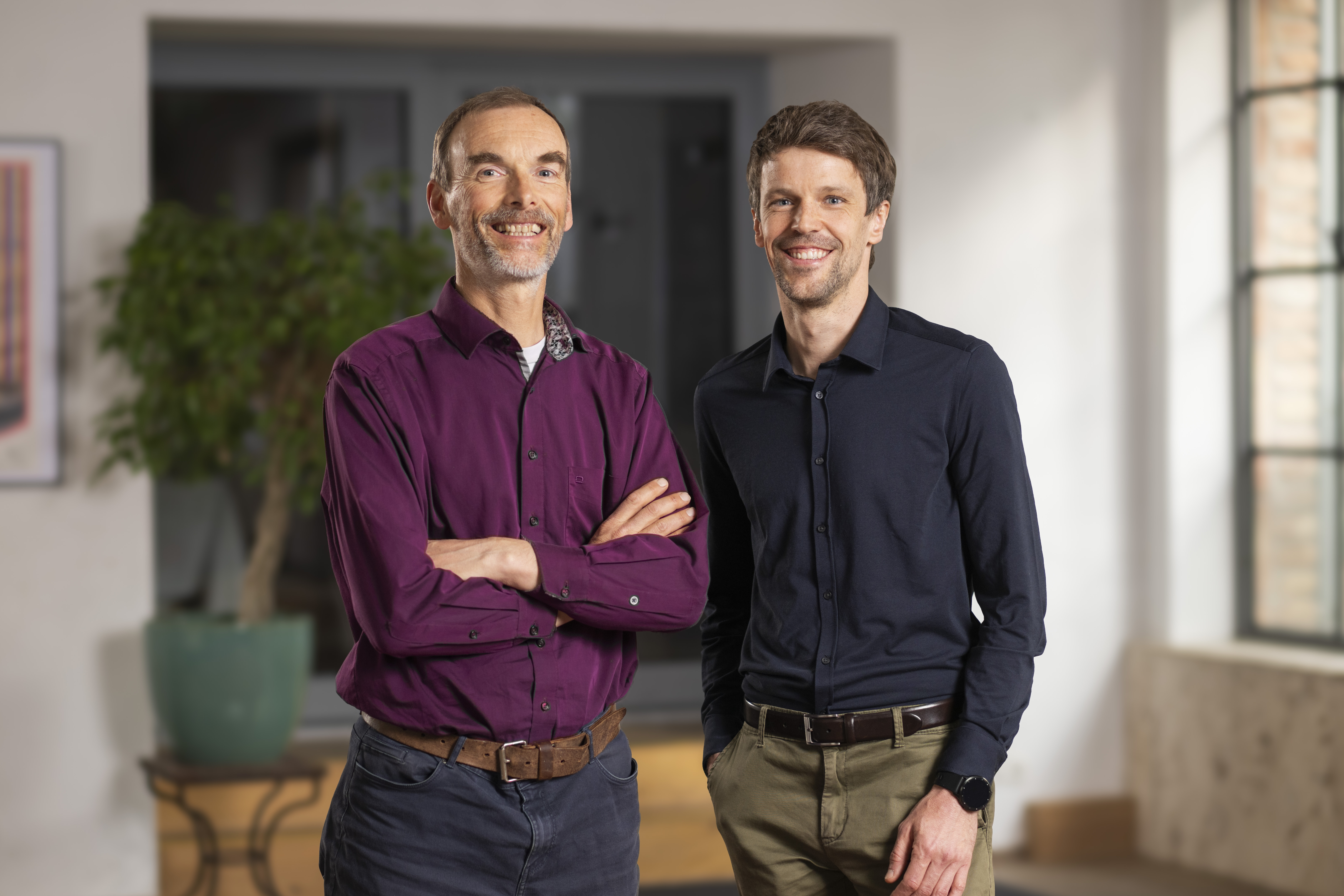 _DSC5120 Zwei Männer stehen in einem Haus, beide lächeln in die Kamera. Einer hat die Arme verschränkt, beide tragen langärmelige Hemden und Hosen. Große Fenster im Hintergrund suggerieren natürliches Licht - perfekt, um über Photovoltaik Funktion zu diskutieren. Eine Topfpflanze sorgt für eine frische Note.