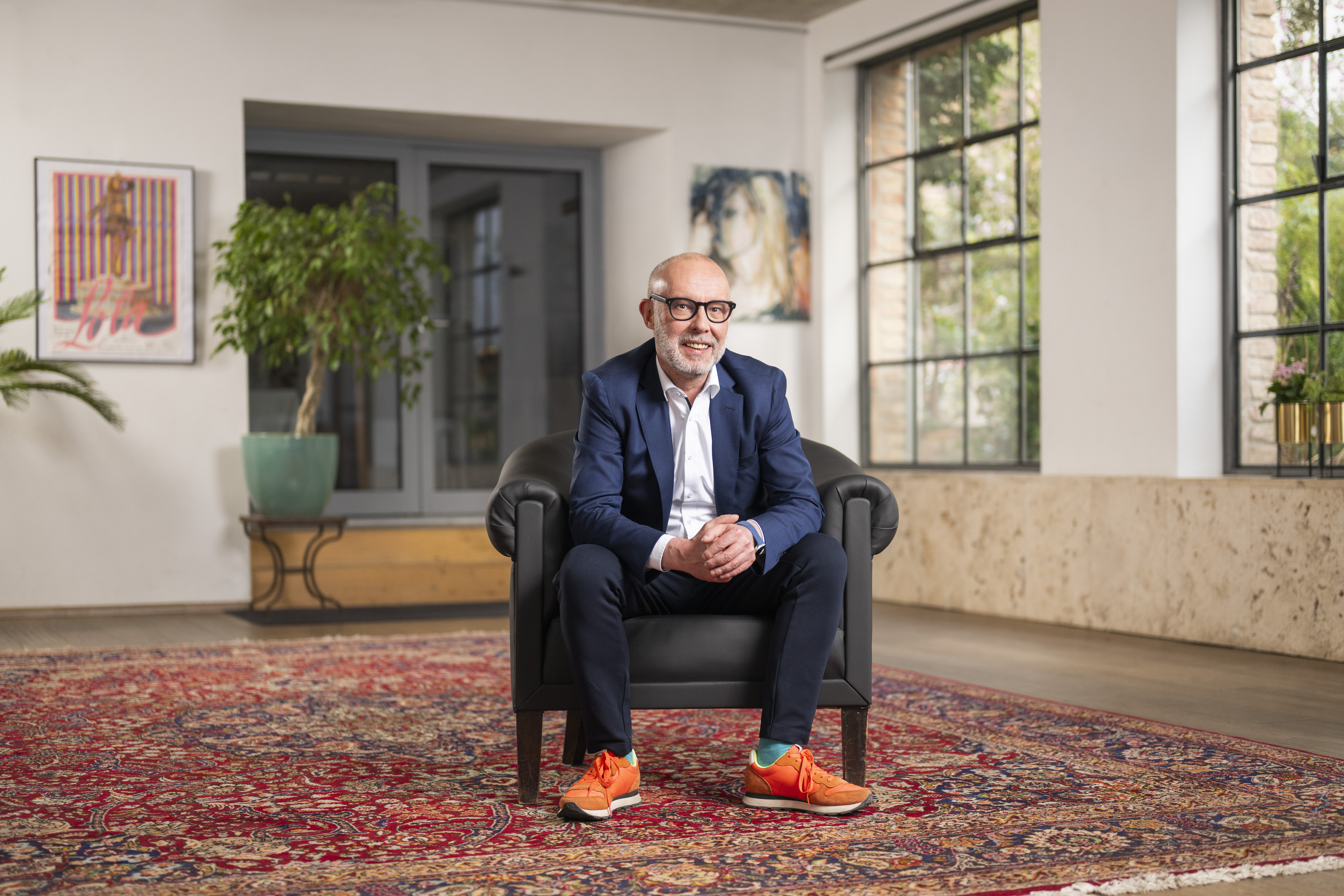 _DSC5284 Ein älterer Mann in einem marineblauen Anzug und leuchtend orangefarbenen Turnschuhen sitzt lächelnd in einem Sessel auf einem bunt gemusterten Teppich im modernen, gut beleuchteten Wohnzimmer des Kraftwerkspark Ökostrom.