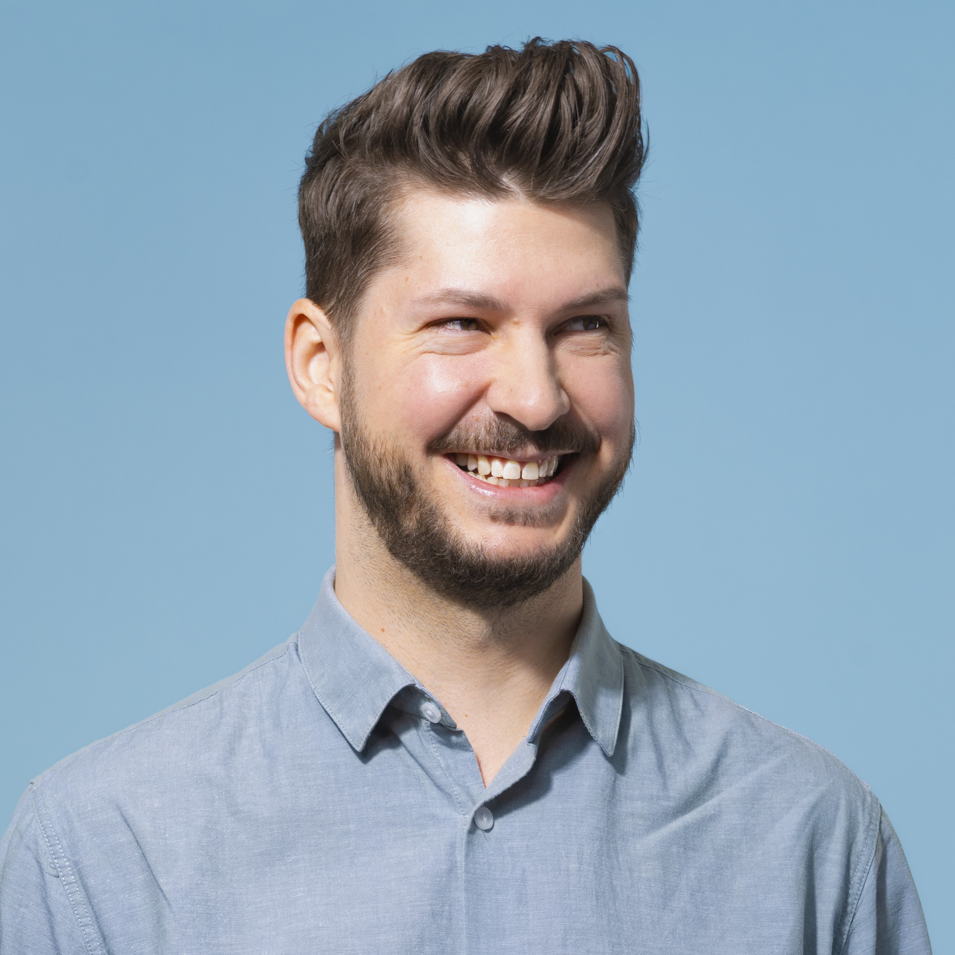 Portrait_ChristianPremm Ein Mann mit kurzen braunen Haaren und Bart, der ein hellblaues Button-up-Hemd trägt, lächelt und blickt nach links vor einem einfarbig blauen Hintergrund.
