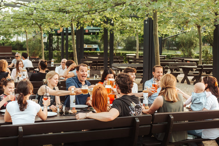 Eine Gruppe von Menschen sitzt an einem langen Tisch im Freien unter einer Pergola, stößt an und unterhält sich in einem Biergarten.
