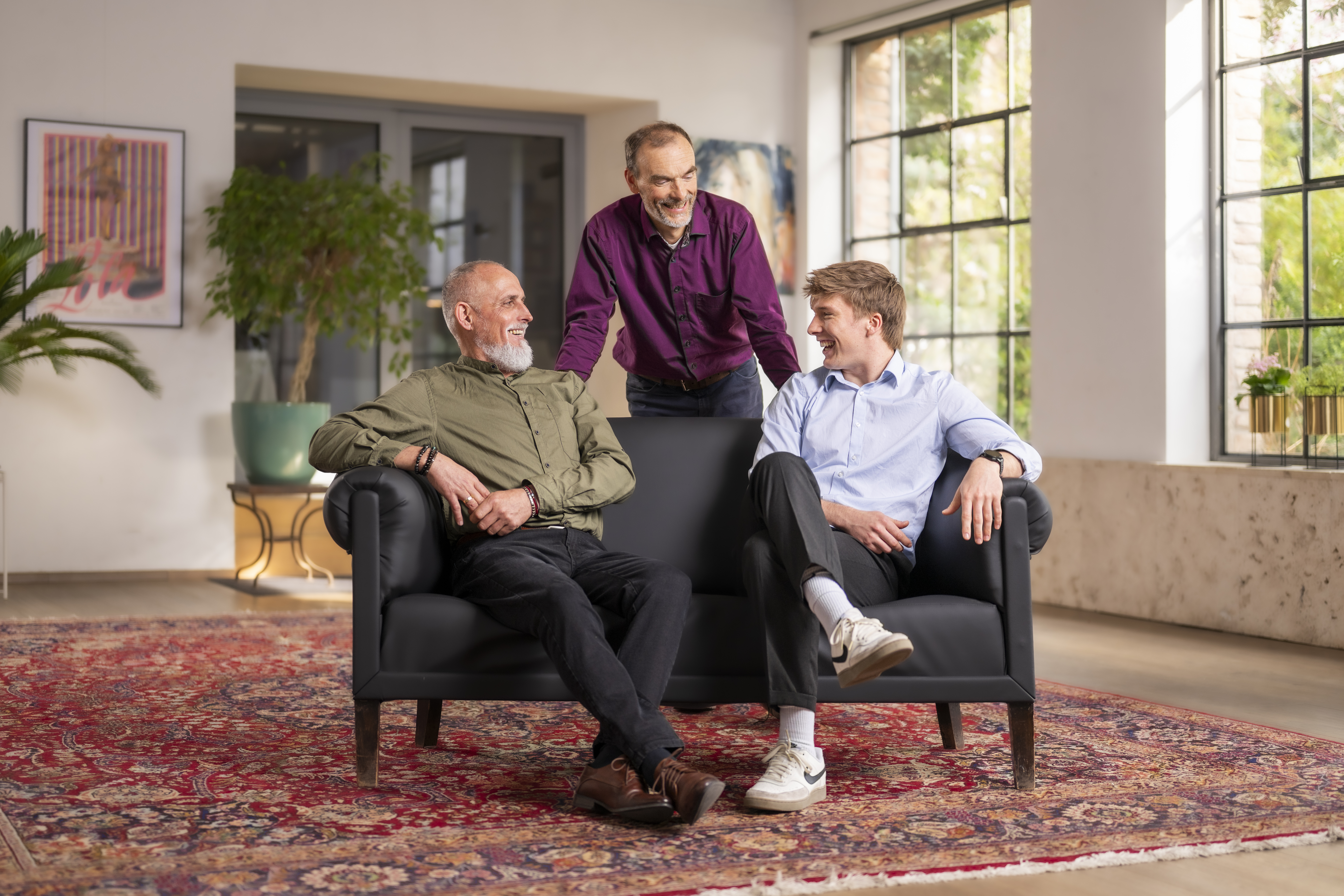_DSC4464 Drei Männer in Freizeitkleidung unterhalten sich und lächeln, während sie in einem Wohnzimmer sitzen und stehen.